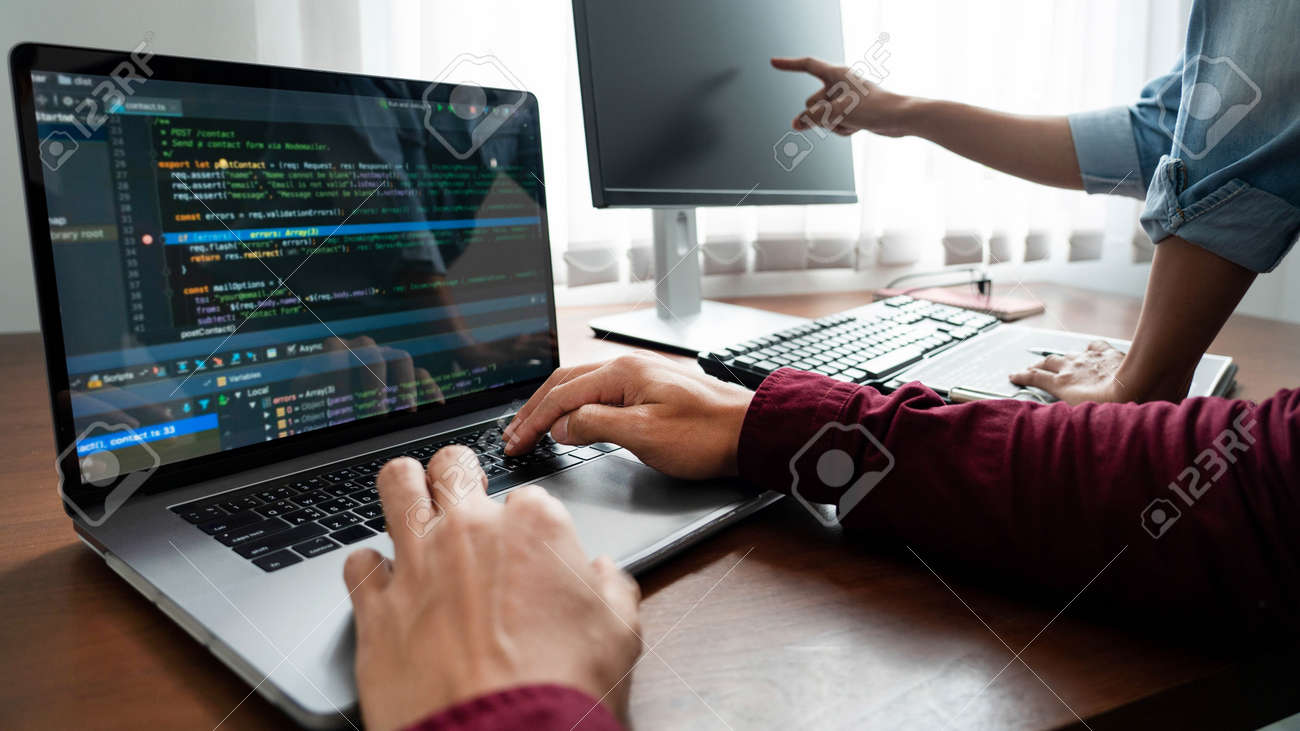 Developer working on laptop at desk with programming code visible and multiple computer screens in collaborative workspace